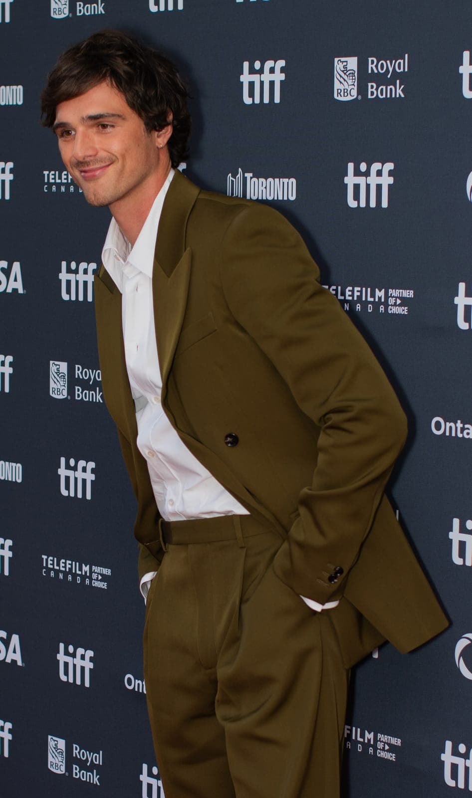 Olive-suit frontal — strong jaw taper, soft smile, dark wave temple control under festival flash.